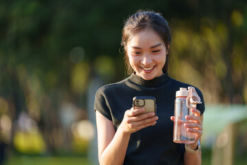 Smiling young asian sportswoman enjoying a break in a park, using her smartphone while holding a water bottle, soaking up the sun after an invigorating training session