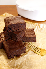Brownies placed with fork on baking paper and blur white bowl at the back with wooden table background. Vertical image of chocolate brownie dessert for homemade, sweet food, and baking snack concepts.