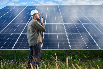 Engineer in uniform with protective helmet near solar station holding walkie talkie. Supervisor is...