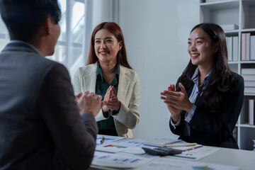 Two women and a man, dressed in professional attire, are clapping and smiling in a modern office setting. Papers and charts are spread on the table, indicating a successful meeting