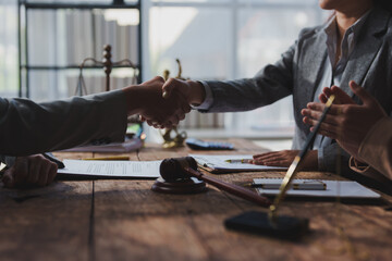 Business professionals shaking hands across a wooden table, symbolizing successful negotiation. Documents, a gavel, and scales of justice are visible, suggesting a legal or corporate setting