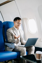 Attractive Asian male passenger of airplane sitting in comfortable seat while working laptop and tablet with mock up area using wireless connection.