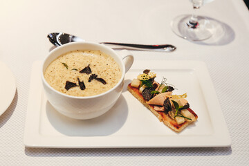 A minimalist shot of a white plate with a serving of French onion soup garnished with truffle slices. Next to it is a gourmet meat bruschetta. The background is clean, with an emphasis on the sophisti