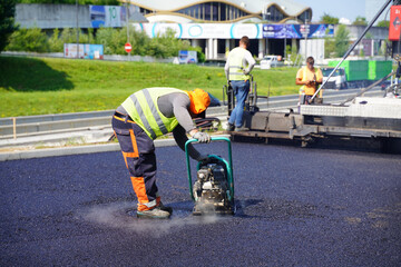 Construction Worker Using Vibratory Plate Compactor on Asphalt with Paver in the Background