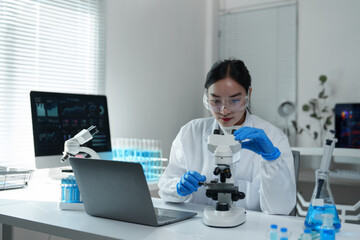 Female scientist wearing lab coat and protective glasses using microscope and laptop, conducting research in modern laboratory with test tubes and computer displaying data analysis