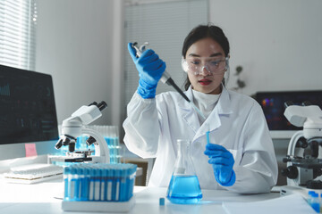 Fototapeta premium Young female scientist wearing lab coat and safety glasses using micropipette dropping blue liquid into laboratory glassware while working on medical research in modern laboratory