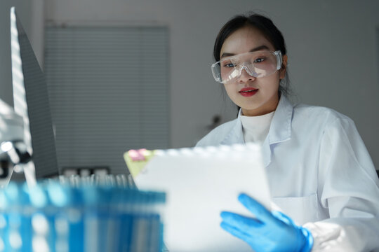 Young female scientist wearing lab coat and protective glasses reading medical research data on clipboard while sitting at desk in modern laboratory