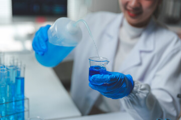 Scientist wearing lab coat and gloves pouring blue liquid reagent from plastic bottle into beaker...