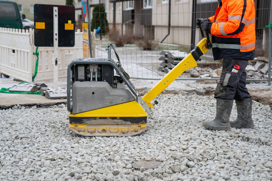 Yellow construction vibratory plate compactor on gravel for ground leveling job site project. Yellow construction equipment. Construction worker wearing vest and working outdoors and cold