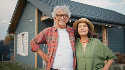 Lovely Caucasian couple standing together. Hugging each other while looking at camera and smiling. One-story building located in background. Proud owners of home in countryside. Village.