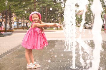 Naklejka premium kid girl in pink dress plays next to fountain on summer day