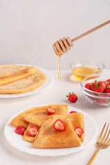Homemade traditional french crepe dish made from unleavened batter with fresh juicy sliced strawberries served on plate with honey pouring from dipper on white wooden table with fork for brunch