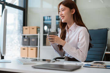 Asian businesswoman sits in a office working on laptop and enjoys a coffee.