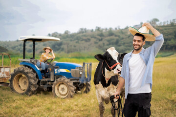 Young Latin man leading cow with smile with blur White woman farmer on tractor background behide.