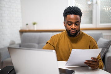 Focused African American businessman using laptop and examining documents while working from his cozy home.	