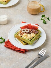 Breakfast, spinach waffles with egg salad, pickled herring and fresh tomato on a white plate on a light concrete background. Waffle recipes.