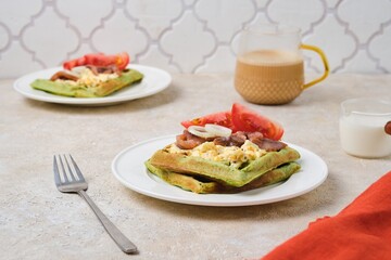 Breakfast, spinach waffles with egg salad, pickled herring and fresh tomato on a white plate on a light concrete background. Waffle recipes.