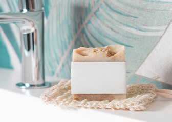 Beige soap with blank label on soap saver bag on basin in green tropical bathroom close up, mockup