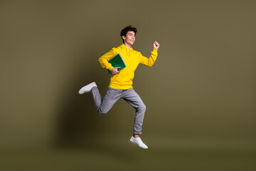 Happy young man jumping with a green book against a solid khaki background in casual yellow sweatshirt