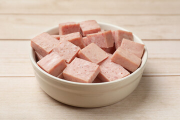 Pieces of canned meat in bowl on white wooden table, closeup