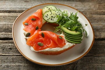 Delicious bagel with salmon, cream cheese, cucumber, avocado and pumpkin seeds on wooden table, closeup