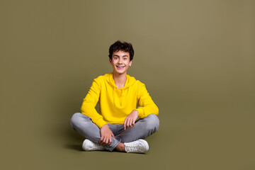 Portrait of a smiling young man in a yellow hoodie, sitting cross-legged against a khaki background
