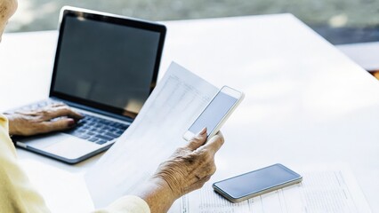 An elderly womans hand sorting through printed health documents and notes surrounded by modern technology illustrating the blend of traditional and digital approaches to managing