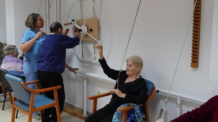 Assisted by nurses, an elderly woman engages in upper body exercises using specialized equipment, promoting mobility and well being within a geriatric care setting