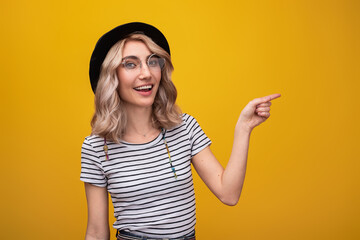 Cheerful woman in casual striped shirt, glasses, and hat pointing with a smile against a vibrant...
