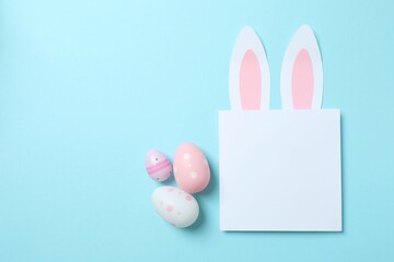 Happy Easter. Colorful eggs and Easter bunny ears with white paper card on a pastel blue background, space for text or advertising