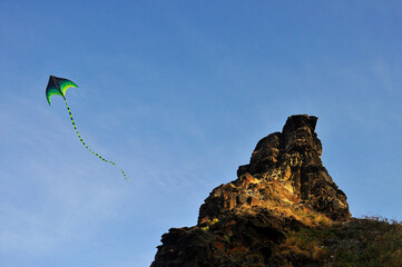 A kite in the blue sky, hovering next to a rock.