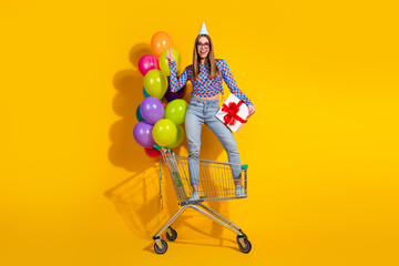 Stylish young woman celebrating with balloons in a shopping cart against a vibrant yellow background