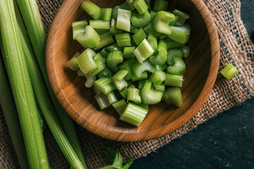 Fresh celery stalks and chopped pieces in a wooden bowl on a rustic dark table. Organic green vegetable on burlap with wooden background. Healthy eating, nutrition, and cooking ingredients concept