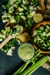 Fresh green smoothie in a textured glass surrounded by chopped ingredients including cucumber, celery, apple, and lemon on a rustic wooden table