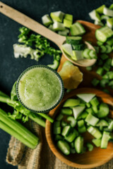 Fresh green smoothie in a textured glass surrounded by chopped ingredients including cucumber,...
