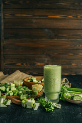 Fresh green smoothie in a textured glass surrounded by chopped ingredients including cucumber,...