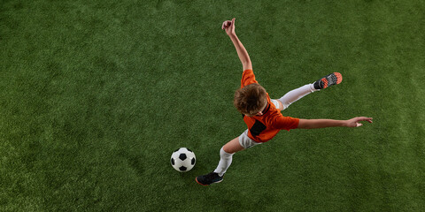 Top view image of boy, soccer player in orange jersey and white shorts in motion of green field, playing, dribbling ball. Training activity. Concept of sport, education, competition, game