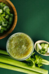 Overhead shot of celery stalks, cucumber, and apple pieces in a wooden bowl, plus a green smoothie on a solid green background. Ideal for healthy eating or detox concepts