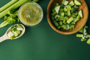 Overhead shot of celery stalks, cucumber, and apple pieces in a wooden bowl, plus a green smoothie on a solid green background. Ideal for healthy eating or detox concepts
