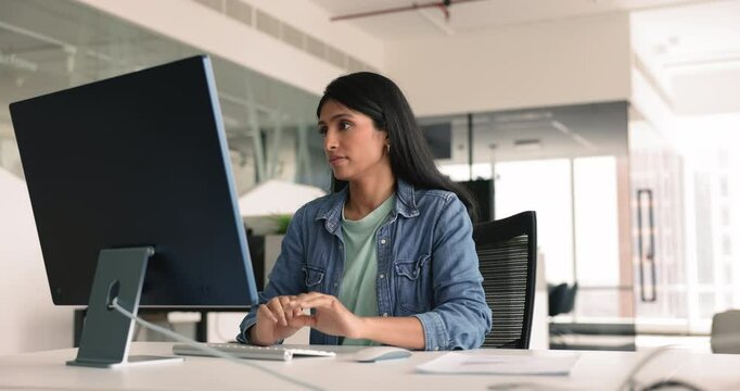 Serious young Indian business professional woman working at computer in office, typing on keyboard, looking at large monitor, testing software product, application, using Internet technology
