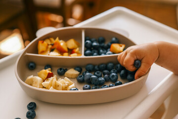 Toddler self-feeding with sliced peaches, bananas, and blueberries from a divided plate on a highchair. Perfect for baby-led weaning, healthy snacking, and developing fine motor skills