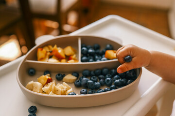 Toddler self-feeding with sliced peaches, bananas, and blueberries from a divided plate on a highchair. Perfect for baby-led weaning, healthy snacking, and developing fine motor skills