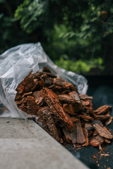 Bag of decorative bark mulch on black landscaping fabric near paving stones and green bushes. Used for soil protection, moisture retention, and weed control in garden landscaping