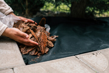 Hands pouring decorative bark mulch onto a black landscape fabric for garden bed protection....