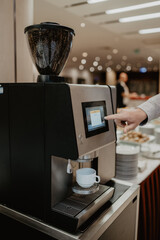 A modern automatic coffee machine with a touchscreen interface dispensing fresh coffee into a white ceramic cup in a well-lit buffet setting