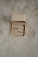 An elegant beige velvet ring box with a pair of wedding bands, including a sleek silver men's band and a delicate diamond-studded women's ring, placed on a soft white faux fur background