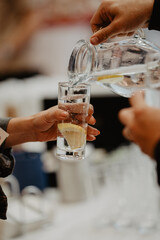A close-up shot of a person pouring fresh water with lemon slices from a glass pitcher into a tall transparent glass, while another person holds the glass,