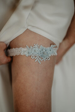  A close-up of a bride lifting her white wedding dress to reveal a delicate lace bridal garter adorned with intricate blue floral embroidery and sparkling rhinestones, symbolizing a timeless wedding 