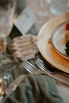 A close-up of a wedding table setting featuring elegantly wrapped guest favors, silver cutlery, a plate with a gourmet dish, and a wine glass, all placed on a soft green fabric table 