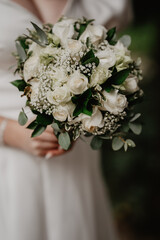 A close-up of a bride holding a beautifully arranged white wedding bouquet featuring elegant roses, delicate baby's breath, and lush green foliage